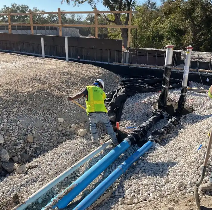 Worker grading gravel around blue water service lines with capped risers on a job site